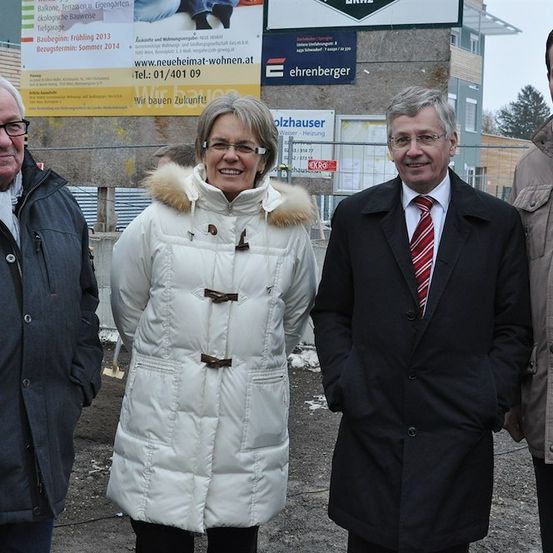 Vier Personen, in Winterkleidung gekleidet, stehen vor einer Baustelle. Die Frau in der Mitte lächelt und trägt einen weißen Pelzmantel. Hinter ihnen befindet sich ein Banner mit Text und Zahlen.