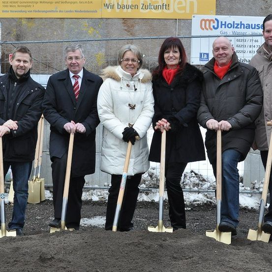 Fünf Personen in formeller Kleidung halten Schaufeln und posieren für ein Foto an einer Baustelle. Hinter ihnen steht ein Schild mit der Aufschrift 'Wir bauen Zukunft!' und weiteren Texten und Zahlen.
