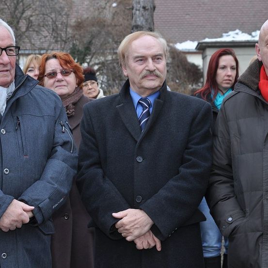 Eine Gruppe von Menschen in Winterkleidung steht vor einem Gebäude mit Schnee auf dem Dach. Der Mann in der Mitte trägt eine Krawatte und einen Schnurrbart.
