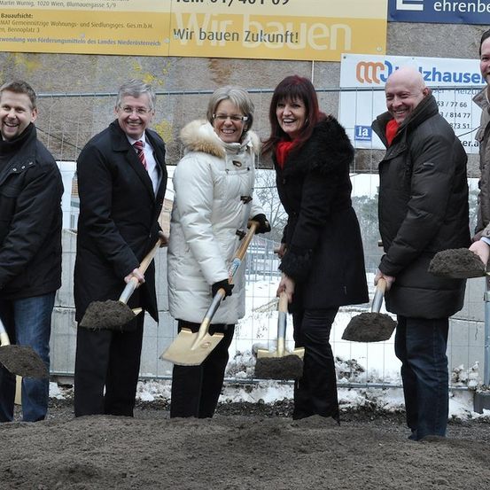 Eine Gruppe von Menschen in Winterkleidung hält Schaufeln und lächelt für ein Foto an einer Baustelle. Hinter ihnen steht ein Banner mit der Aufschrift 'Wir bauen Zukunft!'.