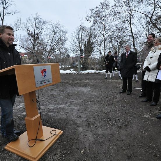 Ein Mann steht an einem Podium mit einem Mikrofon vor einer Gruppe von Menschen auf einem Feld mit Bäumen im Hintergrund.