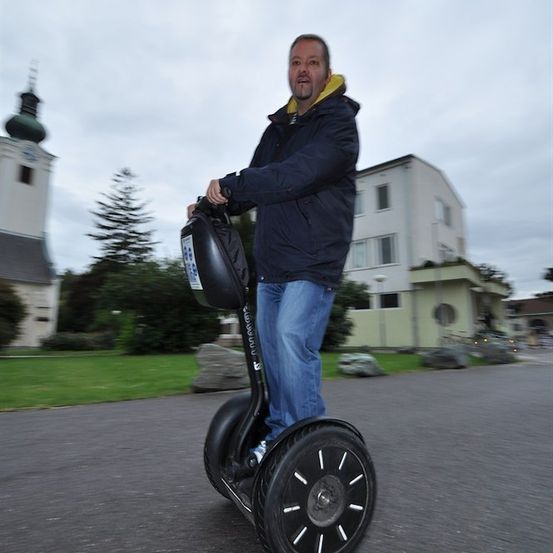 Ein Mann fährt auf einem Segway auf einer asphaltierten Straße, mit einer Kirche und Gebäuden im Hintergrund.