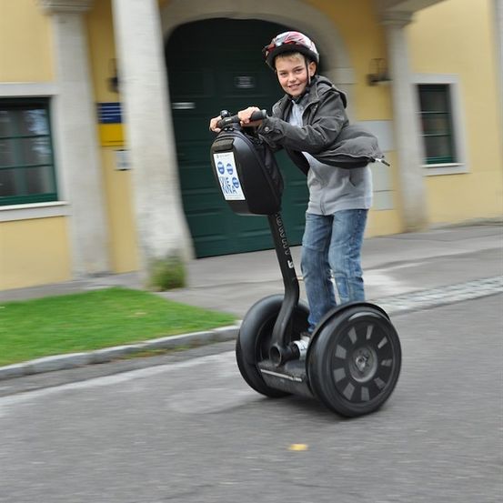Ein junger Junge mit Helm fährt einen Segway auf einer Straße vor einem gelben Gebäude.