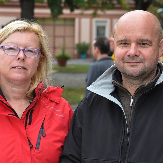 Ein Mann und eine Frau stehen zusammen draußen, die Frau trägt eine Brille und eine rote Jacke, der Mann ist kahlköpfig und trägt eine schwarze Jacke.