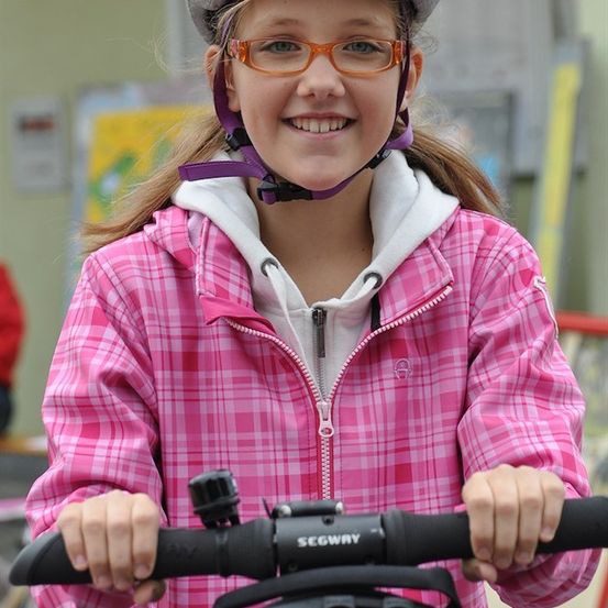 Ein junges Mädchen in einer pinken karierten Jacke und mit Brille lächelt, während es den schwarzen Lenker eines Segways hält.