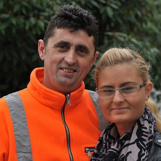 Ein Mann und eine Frau stehen eng beieinander und lächeln für ein Foto. Der Mann trägt eine orange Warnweste und die Frau trägt eine Brille und einen Schal. Sie sind draußen vor einem Baumhintergrund.