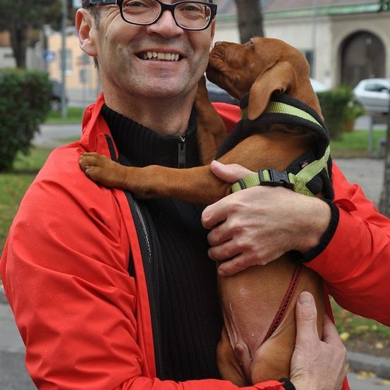Ein Mann mit Brille und roter Jacke hält einen braunen Hund in seinen Armen und lächelt.