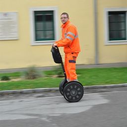 Ein Mann in einem orangefarbenen Sicherheitsanzug fährt auf einer Straße vor einem gelben Gebäude mit einem Segway.