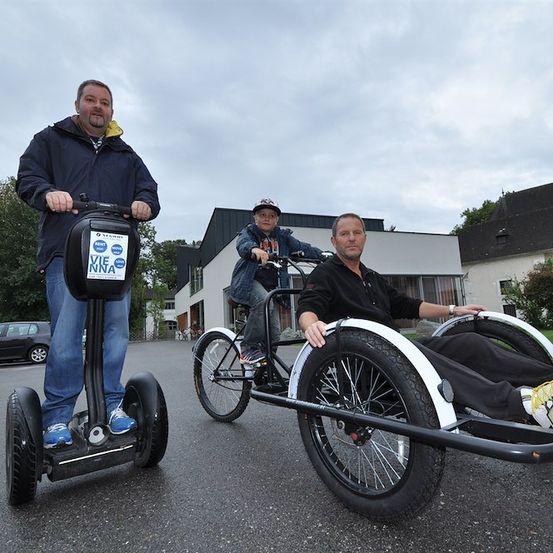Drei Männer sind draußen, zwei fahren einen Segway und ein Dreirad, und einer sitzt auf einem Dreirad. Sie stehen vor einem Gebäude bei bewölktem Himmel.