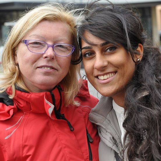 Zwei Frauen stehen eng beieinander, eine mit blondem Haar und Brille, die andere mit langen dunklen Haaren, beide lächeln für ein Foto.