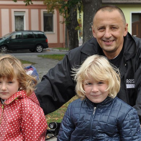 Ein Mann und zwei junge Mädchen stehen vor einem Gebäude mit einem geparkten Auto. Der Mann lächelt und die Mädchen tragen Jacken.