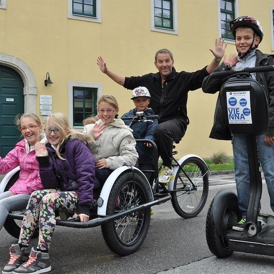Eine Familie mit fünf Personen fährt einen Dreiradwagen, hinter ihnen ein Mann auf einem Segway, alle winken zur Kamera. Der Dreiradwagen hat ein Kind vorne und zwei Kinder hinten. Der Mann auf dem Segway trägt einen Helm.