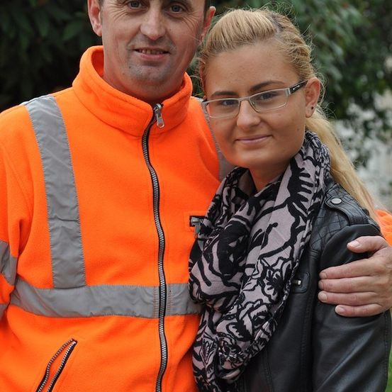 Ein Mann in einer orangefarbenen Sicherheitsjacke und eine Frau in einer schwarzen Lederjacke mit Schal stehen eng beieinander und lächeln für ein Foto.