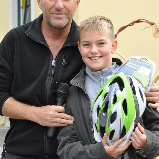 Ein Mann und ein junger Junge stehen zusammen, der Junge hält einen Fahrradhelm und lächelt in die Kamera. Der Mann hält ein Mikrofon und scheint zu sprechen.