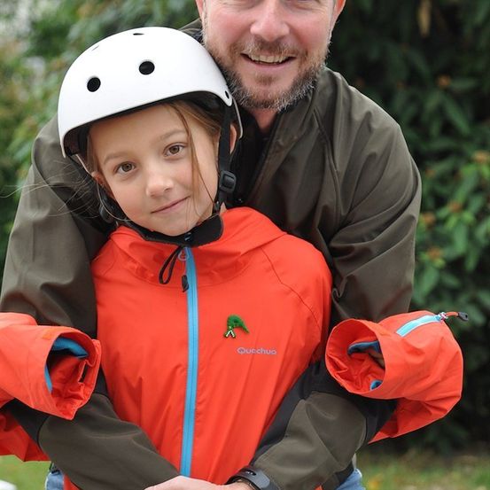 Ein Mann und ein junges Mädchen lächeln und umarmen sich. Das Mädchen trägt eine leuchtend orangefarbene Jacke und einen weißen Helm. Der Mann trägt eine grüne Jacke und hat einen Bart. Sie stehen in einem Grasbereich.