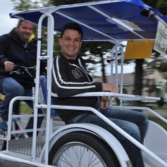 Zwei Männer fahren an einem sonnigen Tag mit einem Dreirad mit blauem Verdeck, einer lächelt und der andere hält das Lenkrad.