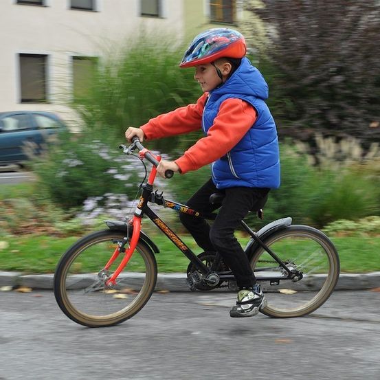 Ein junger Junge mit Helm und blauer Weste fährt ein rotes und schwarzes Fahrrad auf einer Straße.