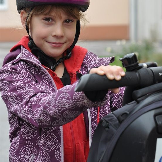 Ein junges Mädchen in einem Helm und bunter Jacke hält das Lenkrad eines schwarzen Rollers.