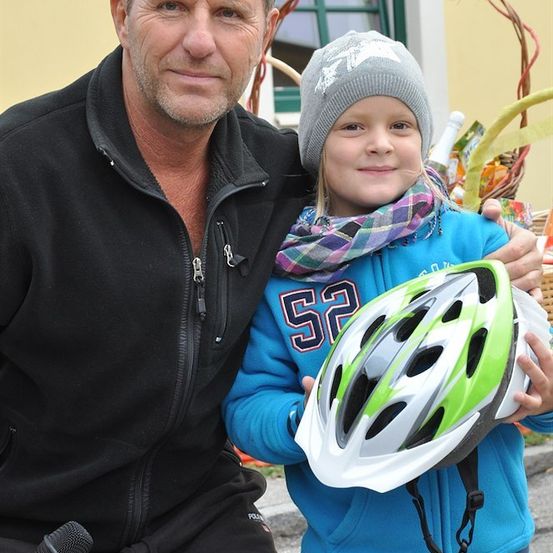 Ein Mann in einem schwarzen Jacken und ein junges Mädchen in einer blauen Jacke und grauer Mütze stehen zusammen. Das Mädchen hält einen grün-weißen Fahrradhelm. Sie lächeln und scheinen für ein Foto zu posieren.