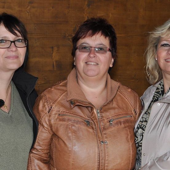 Drei Frauen stehen zusammen vor einer Holzwand und lächeln für ein Foto. Die Frau auf der linken Seite trägt eine Brille und eine Halskette. Die Frau in der Mitte trägt eine braune Lederjacke. Die Frau auf der rechten Seite trägt eine graue Jacke.