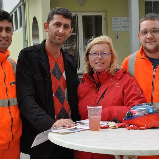 Vier Personen stehen vor einem Gebäude. Zwei Männer tragen orange Westen und eine Frau trägt eine rote Jacke. Ein Mann trägt eine schwarze Jacke und ein rotes Hemd. Ein Helm und ein Becher stehen auf einem Tisch.