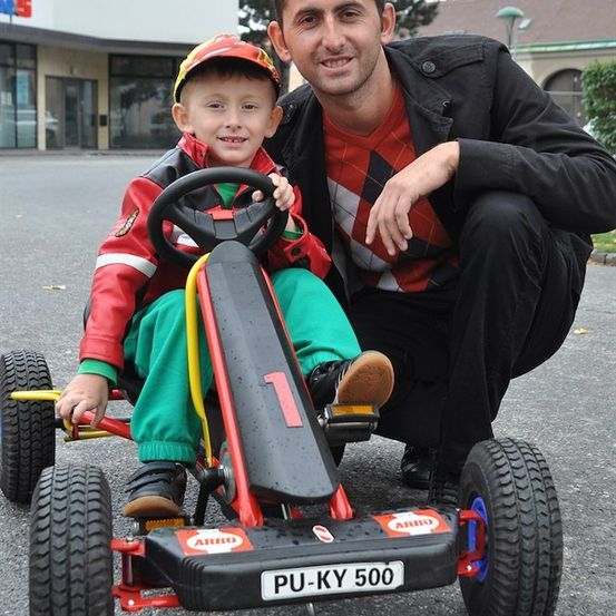 Ein Mann hockt neben einem jungen Jungen in einem Go-Kart, beide lächeln. Der Junge trägt eine rote Jacke und grüne Hose. Der Go-Kart hat ein Nummernschild mit der Aufschrift PU-KY 500.