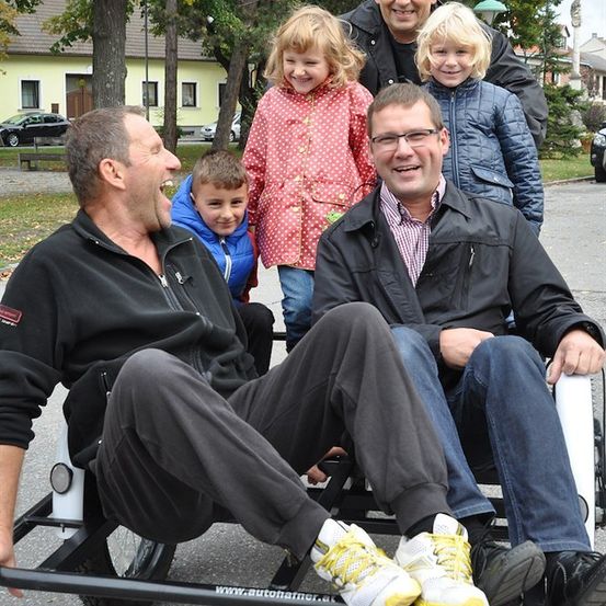 Zwei Männer sitzen auf einem Dreirad, mit drei Kindern dahinter. Die Männer lächeln, und die Kinder stehen vor einem Haus mit Bäumen und geparkten Autos im Hintergrund.