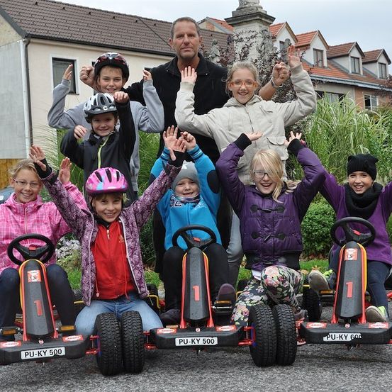 Eine Gruppe von Kindern und Erwachsenen posiert für ein Foto vor einem Haus, alle mit Helmen und lächelnd. Sie sitzen auf Go-Karts mit Nummernschildern.