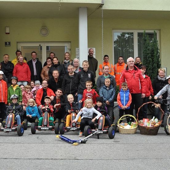 Eine Gruppe von Menschen, einschließlich Kindern, Erwachsenen und einem Hund, posiert für ein Foto vor einem Gebäude mit Körben voller Osterleckerbissen. Einige sitzen auf Go-Karts und Rollern.