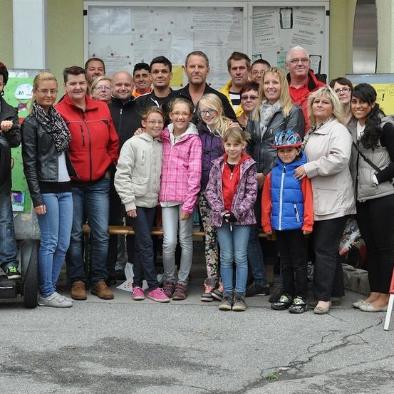 Eine Gruppe von Menschen, darunter Erwachsene und Kinder, posiert für ein Foto vor einem Gebäude mit einem Schild und einem Poster an der Wand. Einige von ihnen tragen Jacken, Brillen und Turnschuhe.