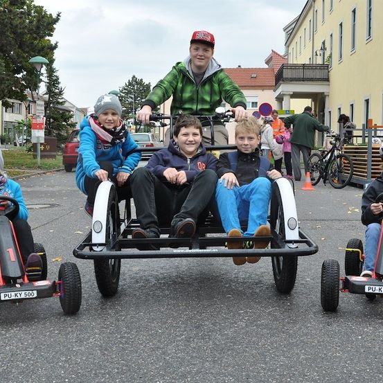 Fünf Kinder fahren auf einem großen Dreirad auf einer Straße. Der Junge in der Mitte trägt einen roten Hut. Hinter ihnen steht ein Mann und hält das Lenkrad. Gebäude und Bäume sind im Hintergrund.