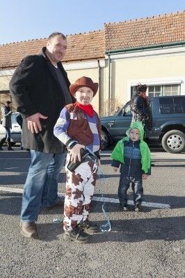 Ein Mann und zwei Kinder in Kostümen stehen auf einem Parkplatz mit einem Gebäude und Autos im Hintergrund.