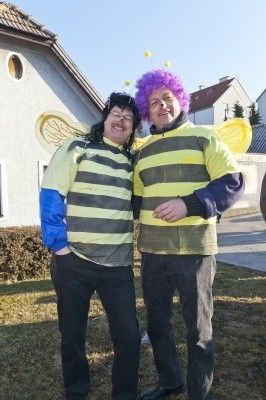 Zwei Menschen in Bienenkostümen stehen nebeneinander auf einem Rasen vor einem Haus.