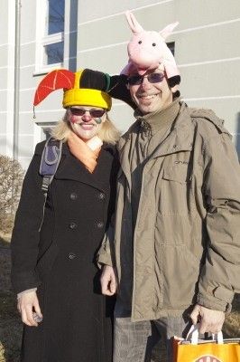 Ein Mann und eine Frau in Kostümen und Hüten, die nebeneinander vor einem Gebäude stehen. Die Frau trägt einen schwarzen Mantel und der Mann einen braunen.