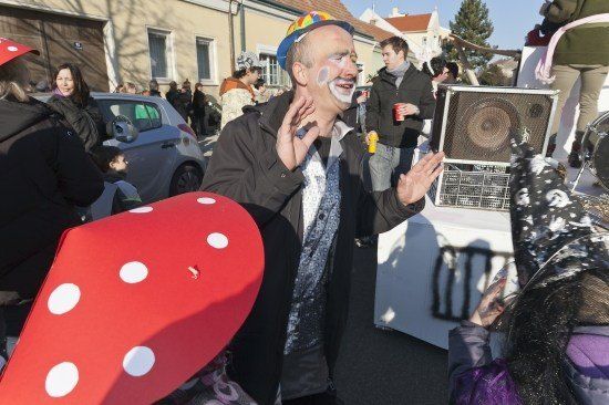 Ein Mann in einem Clownskostüm gestikuliert mit den Händen, während er vor einer Menschenmenge steht. Er ist von Leuten umgeben, von denen einige Kostüme tragen. Im Hintergrund befindet sich ein Gebäude mit einem Baum davor.