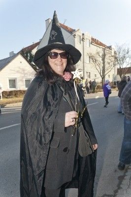 Eine Frau in einer Hexenkostümierung steht auf der Straße, trägt einen schwarzen Hut, Umhang und Sonnenbrille und hält einen Sternenstab. Hinter ihr gehen mehrere Personen, und Gebäude sind sichtbar.
