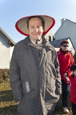 Ein Mann mit einem breitkrempigen Hut und einem grauen Mantel steht auf einer Rasenfläche mit einem Haus und zwei Frauen im Hintergrund.