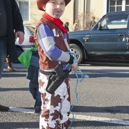 Ein junger Junge, der als Cowboy verkleidet ist, steht auf der Straße und hält eine Seil. Dahinter sind ein Auto und ein Gebäude zu sehen.