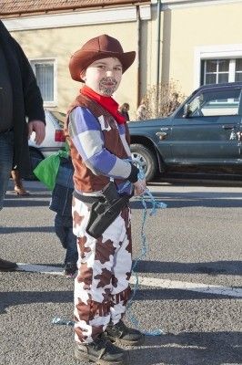 Ein junger Junge, der als Cowboy verkleidet ist, steht auf der Straße und hält eine Seil. Dahinter sind ein Auto und ein Gebäude zu sehen.