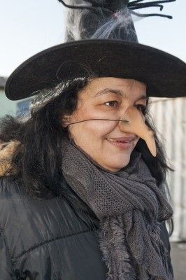 Eine Frau mit einer Hexenmütze und einer langen Nase, lächelt in die Kamera, mit einem Schal um den Hals.