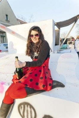 Eine Frau in einem roten Kleid mit Punkten und schwarzen Stiefeln sitzt auf einer weißen Plattform, hält eine Flasche Champagner und lächelt in die Kamera.