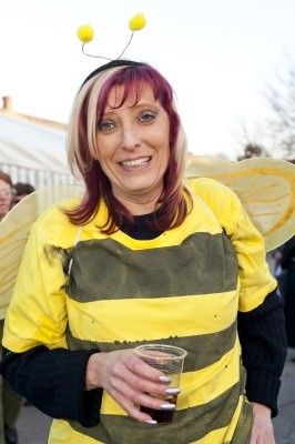 Eine Frau in einem Bienenkostüm, lächelt und hält ein Getränk in einem Plastikbecher, mit einem verschwommenen Hintergrund von Menschen und Bäumen.