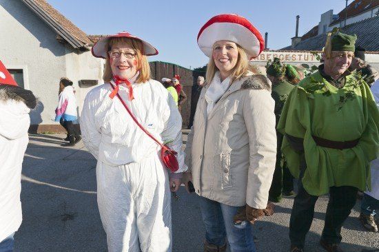 Zwei Frauen in Kostümen, eine in einem weißen Overall und die andere in einem rot-weißen Hut, stehen zusammen mit einem Mann in einem grünen Kostüm mit Blättern. Sie sind draußen, wahrscheinlich bei einem Festival.