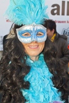 Eine Frau mit langen schwarzen Haaren trägt einen blauen Federschal und eine silberne Maske mit blauen Federn auf dem Kopf und lächelt in die Kamera.
