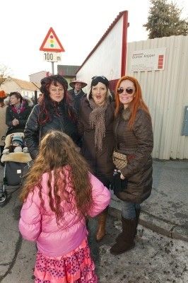 Vier Frauen und ein junges Mädchen stehen draußen, eine Frau hält ein Baby in einem Kinderwagen. Sie sind warm gekleidet und scheinen auf etwas zu warten. Hinter ihnen steht ein Gebäude mit einem Schild und einem Zaun.