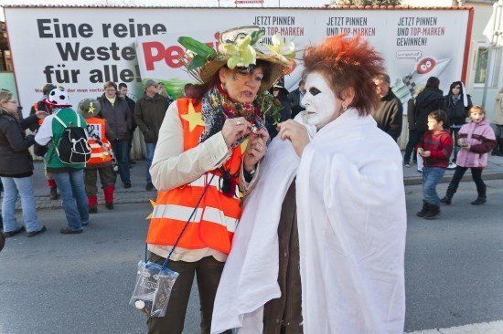 Zwei Menschen in Kostümen stehen auf einer Straße mit einer Menschenmenge im Hintergrund. Einer trägt eine reflektierende Weste und einen Hut mit Blumen, während der andere weißes Gesichtsmake-up und einen weißen Umhang trägt. Ein großes Schild hinter ihnen lautet 'Eine reine Weste für alle!' auf Deutsch.