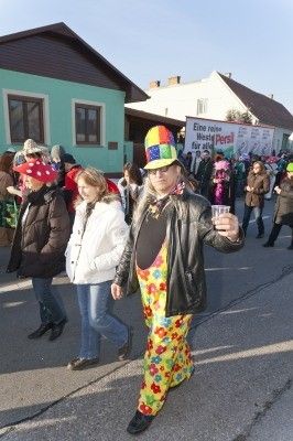 Ein Mann in einem bunten Clownskostüm geht während eines Umzugs die Straße entlang und hält einen Becher. Hinter ihm gehen mehrere Menschen in Winterkleidung in die entgegengesetzte Richtung. Gebäude säumen die Straße.