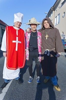 Drei Personen in Kostümen, eine in einem roten Gewand mit einem Kreuz, eine andere in einer rosa Jacke und die dritte in einem braunen Gewand, stehen zusammen auf einer Straße.