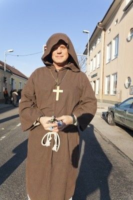 Ein Mann in einem braunen Franziskanergewand steht auf der Straße und hält einen Rosenkranz. Im Hintergrund befinden sich Gebäude und ein geparktes Auto.