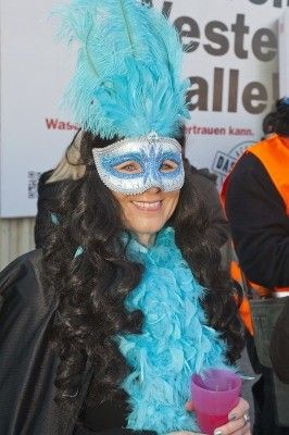 Eine Frau mit langen dunklen Haaren trägt einen blauen Federboa, eine blaue Federhaube und eine silberne Maske. Sie lächelt und hält einen rosafarbenen Becher.